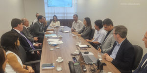 A imagem retrata uma reunião formal em uma sala de conferências. Um grupo de aproximadamente 12 pessoas, incluindo homens e mulheres vestidos com roupas sociais, estão sentados ao redor de uma mesa retangular de madeira. Alguns participantes seguram canetas e fazem anotações, enquanto outros escutam atentamente. Na mesa, há papéis, cadernos, celulares, notebooks e copos de água. Ao fundo, uma tela de projeção exibe um slide com o logotipo do TJMG. O texto na imagem informa que a reunião trata da participação dos sindicatos no Grupo de Trabalho sobre lotação paradigma no TJMG, conforme a Resolução 219 do CNJ.