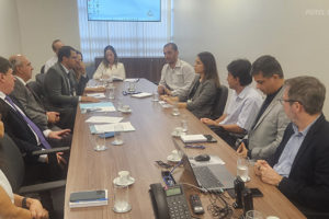 A imagem retrata uma reunião formal em uma sala de conferências. Um grupo de aproximadamente 12 pessoas, incluindo homens e mulheres vestidos com roupas sociais, estão sentados ao redor de uma mesa retangular de madeira. Alguns participantes seguram canetas e fazem anotações, enquanto outros escutam atentamente. Na mesa, há papéis, cadernos, celulares, notebooks e copos de água. Ao fundo, uma tela de projeção exibe um slide com o logotipo do TJMG. O texto na imagem informa que a reunião trata da participação dos sindicatos no Grupo de Trabalho sobre lotação paradigma no TJMG, conforme a Resolução 219 do CNJ.