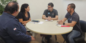 Quatro pessoas estão sentadas ao redor de uma mesa redonda em uma sala de reuniões. Eduardo Amorim (Sindesemp/MG), Luíza Homen (ALMG), Eduardo Couto (Serjusmig) e Alexandre Pires (SINJUS). A mulher, vestindo roupa escura, está falando, enquanto os outros três, usando camisetas polo com logotipos sindicais, escutam atentamente. A parede ao fundo é clara, e há uma planta decorativa no canto.