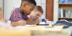 A imagem mostra um ambiente escolar com um menino vestindo uma camisa listrada, concentrado enquanto escreve em um caderno. Ao fundo, uma menina, com óculos na cabeça, também está envolvida em atividades escolares. A mesa está coberta por materiais como cadernos, canetas e lápis. O cenário inclui estantes com livros, sugerindo um ambiente de sala de aula ou biblioteca. A composição da imagem transmite foco e aprendizado.