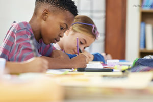 A imagem mostra um ambiente escolar com um menino vestindo uma camisa listrada, concentrado enquanto escreve em um caderno. Ao fundo, uma menina, com óculos na cabeça, também está envolvida em atividades escolares. A mesa está coberta por materiais como cadernos, canetas e lápis. O cenário inclui estantes com livros, sugerindo um ambiente de sala de aula ou biblioteca. A composição da imagem transmite foco e aprendizado.