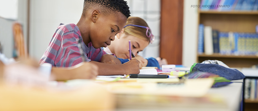 A imagem mostra um ambiente escolar com um menino vestindo uma camisa listrada, concentrado enquanto escreve em um caderno. Ao fundo, uma menina, com óculos na cabeça, também está envolvida em atividades escolares. A mesa está coberta por materiais como cadernos, canetas e lápis. O cenário inclui estantes com livros, sugerindo um ambiente de sala de aula ou biblioteca. A composição da imagem transmite foco e aprendizado.