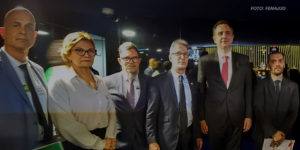 O coordenador-geral do SINJUS, Alexandre Pires, está ao lado do presidente do Senado, Rodrigo Pacheco, e do governador de Minas Gerais, Romeu Zema, na entrada do plenário do Senado. Eles posam para a foto junto com outros participantes do evento, todos vestidos com trajes sociais. O ambiente possui iluminação escura com luzes indiretas ao fundo. A imagem tem o crédito no canto superior direito: "FOTO: FENAJUD"