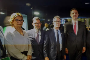 O coordenador-geral do SINJUS, Alexandre Pires, está ao lado do presidente do Senado, Rodrigo Pacheco, e do governador de Minas Gerais, Romeu Zema, na entrada do plenário do Senado. Eles posam para a foto junto com outros participantes do evento, todos vestidos com trajes sociais. O ambiente possui iluminação escura com luzes indiretas ao fundo. A imagem tem o crédito no canto superior direito: "FOTO: FENAJUD"