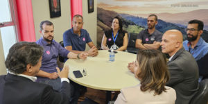 A imagem mostra os dirigentes do SINJUS-MG reunidos com o presidente da FFO, todos ao redor de uma mesa redonda em uma sala de reunião. Alguns usam crachás ou camisetas com adesivos. O encontro trata da aceleração da tramitação do PL da Data-Base 2024, com todos demonstrando atenção e envolvimento na conversa.