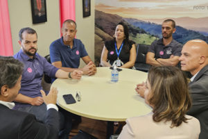 A imagem mostra os dirigentes do SINJUS-MG reunidos com o presidente da FFO, todos ao redor de uma mesa redonda em uma sala de reunião. Alguns usam crachás ou camisetas com adesivos. O encontro trata da aceleração da tramitação do PL da Data-Base 2024, com todos demonstrando atenção e envolvimento na conversa.