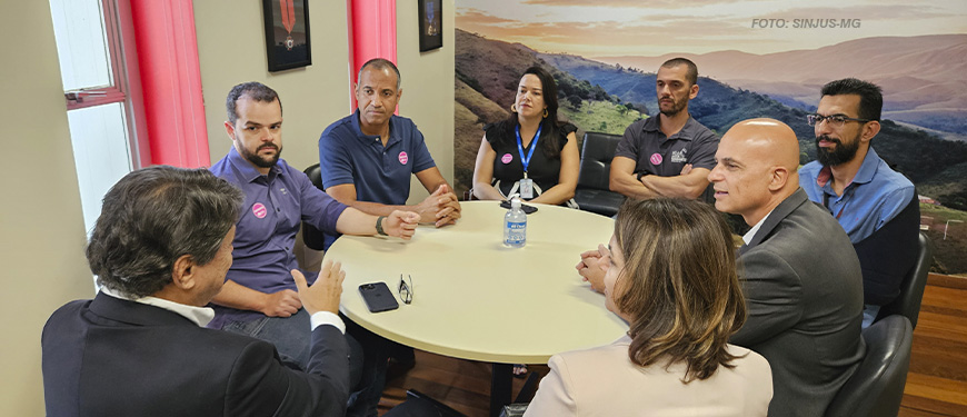 A imagem mostra os dirigentes do SINJUS-MG reunidos com o presidente da FFO, todos ao redor de uma mesa redonda em uma sala de reunião. Alguns usam crachás ou camisetas com adesivos. O encontro trata da aceleração da tramitação do PL da Data-Base 2024, com todos demonstrando atenção e envolvimento na conversa.