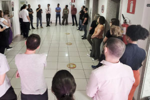 Na foto, observamos uma reunião setorial do SINJUS-MG na unidade Praça Milton Campos do TJMG. Os servidores estão dispostos em círculo em um corredor amplo, demonstrando engajamento coletivo. Eles escutam atentamente as orientações e discussões promovidas por membros da diretoria do sindicato, que se encontram ao fundo, coordenando a conversa.