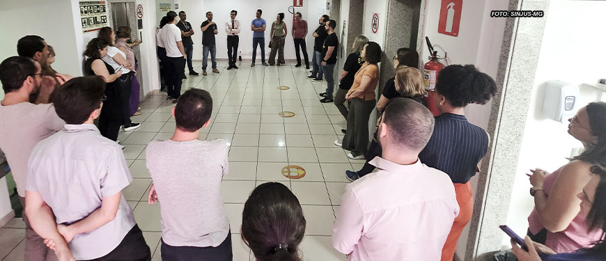 Na foto, observamos uma reunião setorial do SINJUS-MG na unidade Praça Milton Campos do TJMG. Os servidores estão dispostos em círculo em um corredor amplo, demonstrando engajamento coletivo. Eles escutam atentamente as orientações e discussões promovidas por membros da diretoria do sindicato, que se encontram ao fundo, coordenando a conversa.