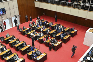 A imagem mostra uma sessão parlamentar em um auditório amplo, com um carpete vermelho e várias mesas e cadeiras organizadas em fileiras. Diversos políticos e servidores estão presentes. Há um telão na parede ao fundo exibindo imagens ao vivo do evento. O destaque é a informação textual: "Luta dos Sindicatos garante aprovação do Orçamento 2025 com redução de danos aos servidores". A atmosfera é formal e demonstra um momento importante de deliberação legislativa.