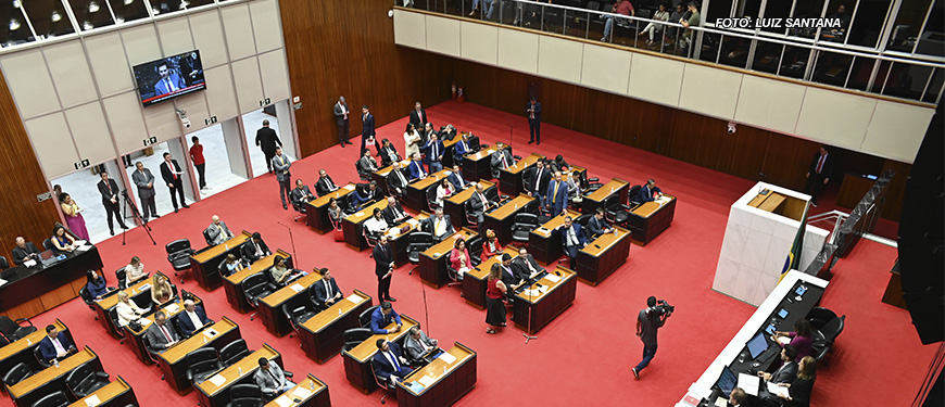 A imagem mostra uma sessão parlamentar em um auditório amplo, com um carpete vermelho e várias mesas e cadeiras organizadas em fileiras. Diversos políticos e servidores estão presentes. Há um telão na parede ao fundo exibindo imagens ao vivo do evento. O destaque é a informação textual: "Luta dos Sindicatos garante aprovação do Orçamento 2025 com redução de danos aos servidores". A atmosfera é formal e demonstra um momento importante de deliberação legislativa.