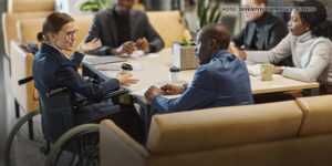 A imagem mostra uma reunião em andamento em um ambiente corporativo. Ao redor de uma mesa estão cinco pessoas, sendo uma delas uma mulher branca em uma cadeira de rodas, que parece estar liderando a conversa, gesticulando enquanto fala. Os demais participantes prestam atenção ou interagem com ela, com expressões sérias e interessadas. No fundo, há móveis modernos e plantas decorativas, conferindo um ar profissional e acolhedor ao espaço.