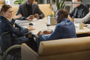 A imagem mostra uma reunião em andamento em um ambiente corporativo. Ao redor de uma mesa estão cinco pessoas, sendo uma delas uma mulher branca em uma cadeira de rodas, que parece estar liderando a conversa, gesticulando enquanto fala. Os demais participantes prestam atenção ou interagem com ela, com expressões sérias e interessadas. No fundo, há móveis modernos e plantas decorativas, conferindo um ar profissional e acolhedor ao espaço.