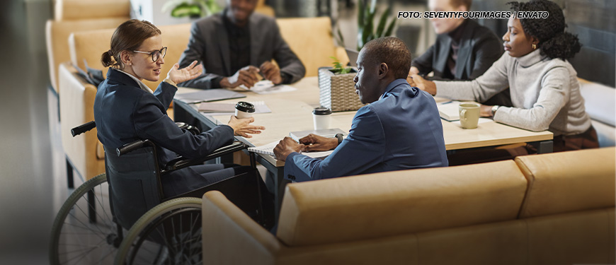 A imagem mostra uma reunião em andamento em um ambiente corporativo. Ao redor de uma mesa estão cinco pessoas, sendo uma delas uma mulher branca em uma cadeira de rodas, que parece estar liderando a conversa, gesticulando enquanto fala. Os demais participantes prestam atenção ou interagem com ela, com expressões sérias e interessadas. No fundo, há móveis modernos e plantas decorativas, conferindo um ar profissional e acolhedor ao espaço.