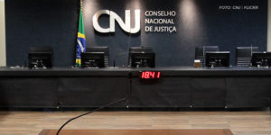 Imagem Acessível: Mesa diretiva do Conselho Nacional de Justiça, ao fundo está um letreiro em metal prateado brilhante com as iniciais CNJ ocupando o centro da imagem, ao lado da sigla está a bandeira do Brasil. Conteúdo textual: Cotas raciais - Fenajud representa no CNJ contra Desembargadora do TJBA por declarações preconceituosas.