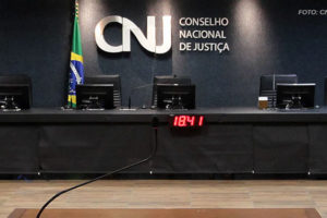 Imagem Acessível: Mesa diretiva do Conselho Nacional de Justiça, ao fundo está um letreiro em metal prateado brilhante com as iniciais CNJ ocupando o centro da imagem, ao lado da sigla está a bandeira do Brasil. Conteúdo textual: Cotas raciais - Fenajud representa no CNJ contra Desembargadora do TJBA por declarações preconceituosas.