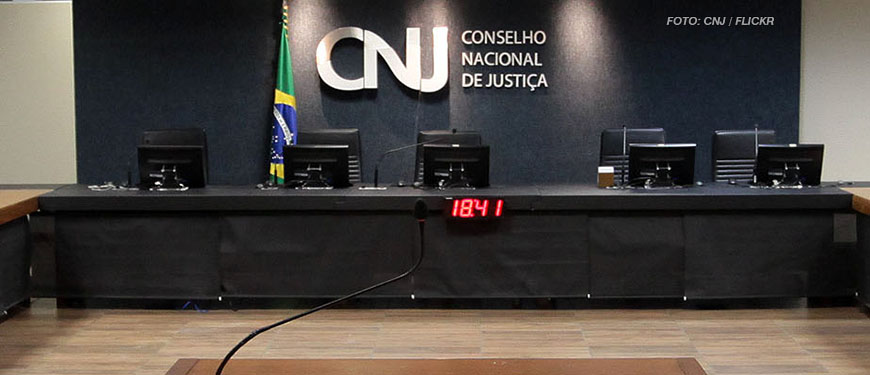 Imagem Acessível: Mesa diretiva do Conselho Nacional de Justiça, ao fundo está um letreiro em metal prateado brilhante com as iniciais CNJ ocupando o centro da imagem, ao lado da sigla está a bandeira do Brasil. Conteúdo textual: Cotas raciais - Fenajud representa no CNJ contra Desembargadora do TJBA por declarações preconceituosas.