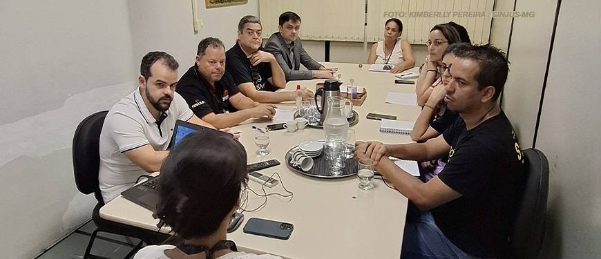 A imagem retrata uma reunião com a DEARHU, na qual várias pessoas estão sentadas ao redor de uma mesa em um ambiente formal. No centro da mesa, há jarros de água, copos e utensílios, sugerindo um encontro organizado. Alguns participantes utilizam computadores abertos e possuem materiais de anotação, indicando uma discussão focada no esclarecimento de informações nos contracheques. Na parte superior da imagem, há um texto que enfatiza a busca dos sindicatos por maior clareza nos contracheques do Tribunal de Justiça de Minas Gerais (TJMG).