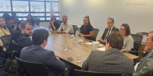 As imagens mostram uma reunião formal com os integrantes do SINJUS, do SERJUSMIG, do TJMMG e da AGE em uma sala de conferências iluminada, com uma grande mesa retangular. Os participantes estão sentados e escutam atentamente. Há copos de água, celulares e documentos sobre a mesa. No fundo, grandes janelas revelam uma vista urbana.