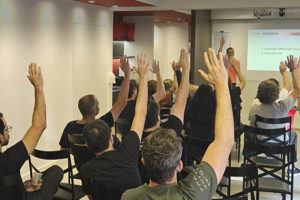 A imagem mostra uma sala de conferências do SINJUS-MG, onde há vários servidores sentados e assistindo a apresentação da pauta de reivindicações, à frente se vê o coordenador geral, Alexandre Paulo Pires da Silva que está direcionando as pautas.