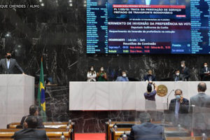 Deputados estaduais conversando na Mesa Diretora e no Plenário da Assembleia Legislativa de Minas Gerais. Ao fundo há uma parede em granito preto e um grande telão que projeta a imagem de um parlamentar que discursa na tribuna.
