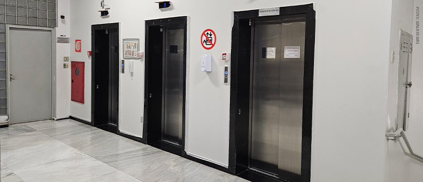 A imagem mostra um corredor do Anexo 1, com quatro elevadores lado a lado, todos de portas metálicas e moldura preta. Acima de cada elevador há um visor digital azul indicando o andar em que cada cabine está.