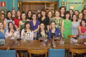 As imagens mostram a confraternização do Núcleo das Mulheres, onde um grupo diverso de mulheres celebra um ciclo em um restaurante, sentadas em mesas compridas e alegres, segurando taças de vinho. Elas aparecem bem arrumadas, sorrindo e interagindo entre si. O ambiente tem uma decoração acolhedora e vibrante, com detalhes coloridos nas paredes. A atmosfera é festiva, destacando a união e o espírito colaborativo do grupo.