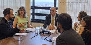 A imagem mostra a reunião com 1° vice do TJ em uma sala de conferências. Seis pessoas estão sentadas ao redor de uma mesa retangular, conversando e discutindo questões aparentes. Há copos d'água, papéis e canetas sobre a mesa, além de um ambiente bem iluminado por janelas ao fundo, que exibem parte de uma paisagem urbana. A legenda da imagem menciona que o SINJUS está discutindo a terceirização relacionada aos taquígrafos em reunião com o primeiro vice-presidente do Tribunal de Justiça.