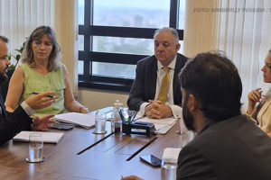 A imagem mostra a reunião com 1° vice do TJ em uma sala de conferências. Seis pessoas estão sentadas ao redor de uma mesa retangular, conversando e discutindo questões aparentes. Há copos d'água, papéis e canetas sobre a mesa, além de um ambiente bem iluminado por janelas ao fundo, que exibem parte de uma paisagem urbana. A legenda da imagem menciona que o SINJUS está discutindo a terceirização relacionada aos taquígrafos em reunião com o primeiro vice-presidente do Tribunal de Justiça.