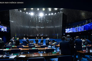 Senadores no Plenário do Senado Federal. Bancadas de madeira em destaque na imagem com cadeiras e carpete em azul. Ao fundo da imagem está a Mesa Diretora.