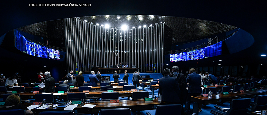 Senadores no Plenário do Senado Federal. Bancadas de madeira em destaque na imagem com cadeiras e carpete em azul. Ao fundo da imagem está a Mesa Diretora.