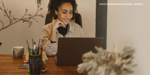 Uma mulher negra e cabelos cacheados presos em um coque alto está sentada à mesa, sorrindo enquanto olha para a tela de seu laptop. Ela usa uma camisa bege sobre uma blusa preta e um colar prateado. Sua mão esquerda apoia o rosto, e sua expressão transmite concentração e satisfação. A mesa de madeira tem um copo com lápis e canetas, um caderno e uma xícara de chá com um desenho de folhas. O ambiente tem uma decoração aconchegante, com plantas e galhos secos ao fundo. Conteúdo textual: PLANEJAMENTO - SINJUS solicita teletrabalho para servidores do TJMG na sexta-feira de Carnaval