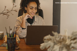 Uma mulher negra e cabelos cacheados presos em um coque alto está sentada à mesa, sorrindo enquanto olha para a tela de seu laptop. Ela usa uma camisa bege sobre uma blusa preta e um colar prateado. Sua mão esquerda apoia o rosto, e sua expressão transmite concentração e satisfação. A mesa de madeira tem um copo com lápis e canetas, um caderno e uma xícara de chá com um desenho de folhas. O ambiente tem uma decoração aconchegante, com plantas e galhos secos ao fundo. Conteúdo textual: PLANEJAMENTO - SINJUS solicita teletrabalho para servidores do TJMG na sexta-feira de Carnaval