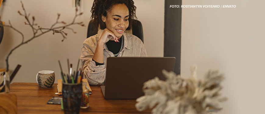 Uma mulher negra e cabelos cacheados presos em um coque alto está sentada à mesa, sorrindo enquanto olha para a tela de seu laptop. Ela usa uma camisa bege sobre uma blusa preta e um colar prateado. Sua mão esquerda apoia o rosto, e sua expressão transmite concentração e satisfação. A mesa de madeira tem um copo com lápis e canetas, um caderno e uma xícara de chá com um desenho de folhas. O ambiente tem uma decoração aconchegante, com plantas e galhos secos ao fundo. Conteúdo textual: PLANEJAMENTO - SINJUS solicita teletrabalho para servidores do TJMG na sexta-feira de Carnaval