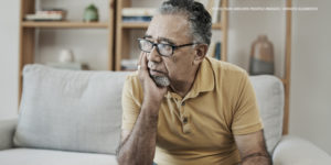 Imagem Acessível: A imagem mostra um homem idoso sentado em um sofá, com uma expressão de preocupação. Ele está usando óculos e uma camisa polo amarela. Ele apoia o rosto em uma das mãos, com o olhar voltado para o lado, parecendo pensativo. Ao fundo, há uma estante com alguns objetos de decoração e livros, sugerindo um ambiente doméstico. Conteúdo textual: “Jabuti” - Reforma da previdência para servidores estaduais e municipais é escamoteada na PEC 66/2023.