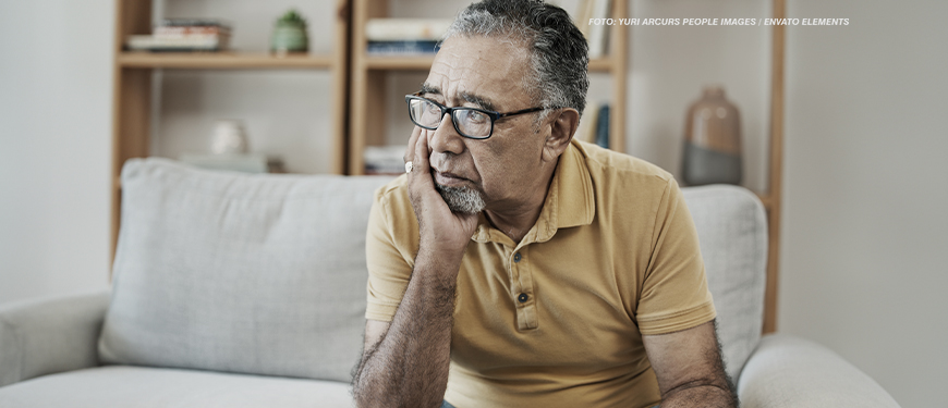 Imagem Acessível: A imagem mostra um homem idoso sentado em um sofá, com uma expressão de preocupação. Ele está usando óculos e uma camisa polo amarela. Ele apoia o rosto em uma das mãos, com o olhar voltado para o lado, parecendo pensativo. Ao fundo, há uma estante com alguns objetos de decoração e livros, sugerindo um ambiente doméstico. Conteúdo textual: “Jabuti” - Reforma da previdência para servidores estaduais e municipais é escamoteada na PEC 66/2023.