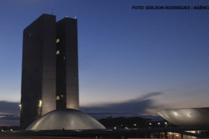 A imagem mostra o prédio do Congresso Nacional em Brasília ao entardecer, com o céu azul escuro ao fundo e luzes acesas nas estruturas. No topo da imagem, há o crédito da foto, mencionando "Edilson Rodrigues/Agência Senado".