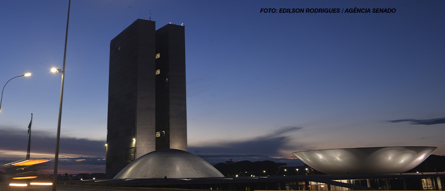 A imagem mostra o prédio do Congresso Nacional em Brasília ao entardecer, com o céu azul escuro ao fundo e luzes acesas nas estruturas. No topo da imagem, há o crédito da foto, mencionando "Edilson Rodrigues/Agência Senado".