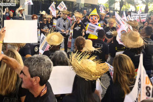 A foto mostra uma manifestação com a temática junina organizada pelos sindicatos SINJUS e SERJUSMIG na frente da sede do TJMG. Várias pessoas estão reunidas, muitas delas vestidas com trajes típicos de festa junina, como chapéus de palha. Alguns manifestantes seguram cartazes e bandeiras. Ao fundo, há uma personagem remetendo ao Pinóquio.