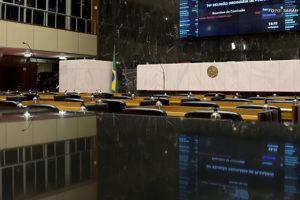 Foto do plenário da Assembleia Legislativa de Minas Gerais (ALMG) com as cadeiras e o púlpito em destaque. Conteúdo textual: ALERTA! - RRF de volta à pauta da ALMG nesta quarta, dia 28, às 17h. Venha lutar contra o projeto.