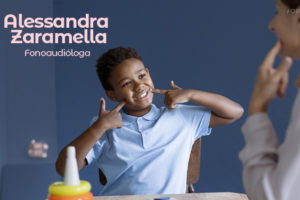 Imagem Acessível: Um menino está sentado em uma mesa, puxando os cantos da boca com os dedos, como se estivesse sorrindo, sugerindo um exercício de fonoaudiologia. Ele veste uma camisa polo azul claro. De frente para ele, está uma mulher de perfil, possivelmente uma fonoaudióloga, observando e participando da atividade. Sobre a imagem há um texto: Alessandra Zaramella - Fonoaudióloga. Conteúdo textual: Fonoaudiologia - Cuide da sua voz com o novo convênio do SINJUS.