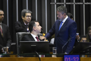 As imagens retratam uma reunião formal no ambiente legislativo brasileiro, com foco na aprovação do Propag. Nelas, há dois homens em destaque, ambos de terno, com um deles tocando o ombro do outro. O fundo apresenta outras pessoas, com uma atmosfera de debate