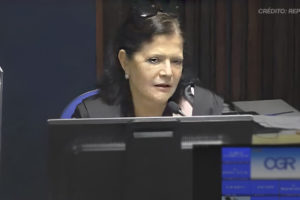 A imagem mostra a desembargadora Rosita Falcão sentada em um ambiente formal, com microfone à sua frente e um monitor em destaque. Ela tem cabelos pretos e usa um traje formal preto. No contexto, há o título: "Fenajud repudia declarações racistas da desembargadora Rosita Falcão sobre cotas raciais", destacando uma nota pública de repúdio sobre comentários racistas.