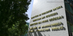 A imagem mostra a fachada do edifício do Ministério da Igualdade Racial. A iluminação natural projeta sombras das letras na parede. No topo da imagem, há um texto sobreposto informando que o Ministério da Igualdade Racial lançou um edital para aprimorar denúncias de racismo.