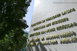 A imagem mostra a fachada do edifício do Ministério da Igualdade Racial. A iluminação natural projeta sombras das letras na parede. No topo da imagem, há um texto sobreposto informando que o Ministério da Igualdade Racial lançou um edital para aprimorar denúncias de racismo.