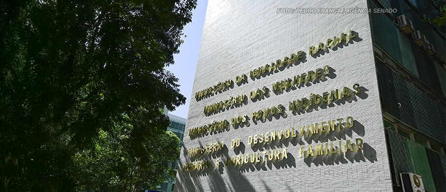 A imagem mostra a fachada do edifício do Ministério da Igualdade Racial. A iluminação natural projeta sombras das letras na parede. No topo da imagem, há um texto sobreposto informando que o Ministério da Igualdade Racial lançou um edital para aprimorar denúncias de racismo.