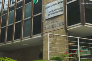 A imagem mostra a fachada de um edifício identificado como "Edifício Presidente Tancredo Neves", com uma placa que indica ser parte do Poder Judiciário, Justiça Militar de Minas Gerais. A placa está localizada à direita da entrada principal do edifício. No topo da placa, há uma bandeira do Brasil parcialmente visível. As janelas do prédio são grandes e retangulares.