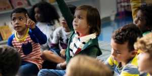 Um grupo de crianças está sentado no chão de uma sala de aula, com algumas levantando as mãos, como se estivessem participando de uma atividade ou respondendo a perguntas. O ambiente sugere uma atmosfera educativa e inclusiva, com prateleiras cheias de livros ao fundo.