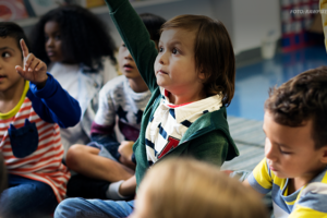 Um grupo de crianças está sentado no chão de uma sala de aula, com algumas levantando as mãos, como se estivessem participando de uma atividade ou respondendo a perguntas. O ambiente sugere uma atmosfera educativa e inclusiva, com prateleiras cheias de livros ao fundo.