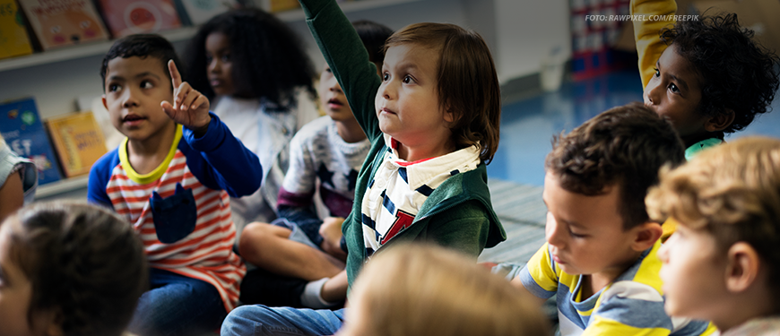 Um grupo de crianças está sentado no chão de uma sala de aula, com algumas levantando as mãos, como se estivessem participando de uma atividade ou respondendo a perguntas. O ambiente sugere uma atmosfera educativa e inclusiva, com prateleiras cheias de livros ao fundo.